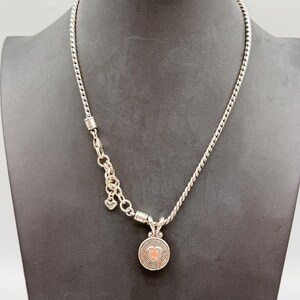 May include: A silver-toned necklace featuring a twisted chain and a circular pendant with a textured geometric pattern and a small heart-shaped center. The necklace includes a short section of larger link chain with a small dangling charm.