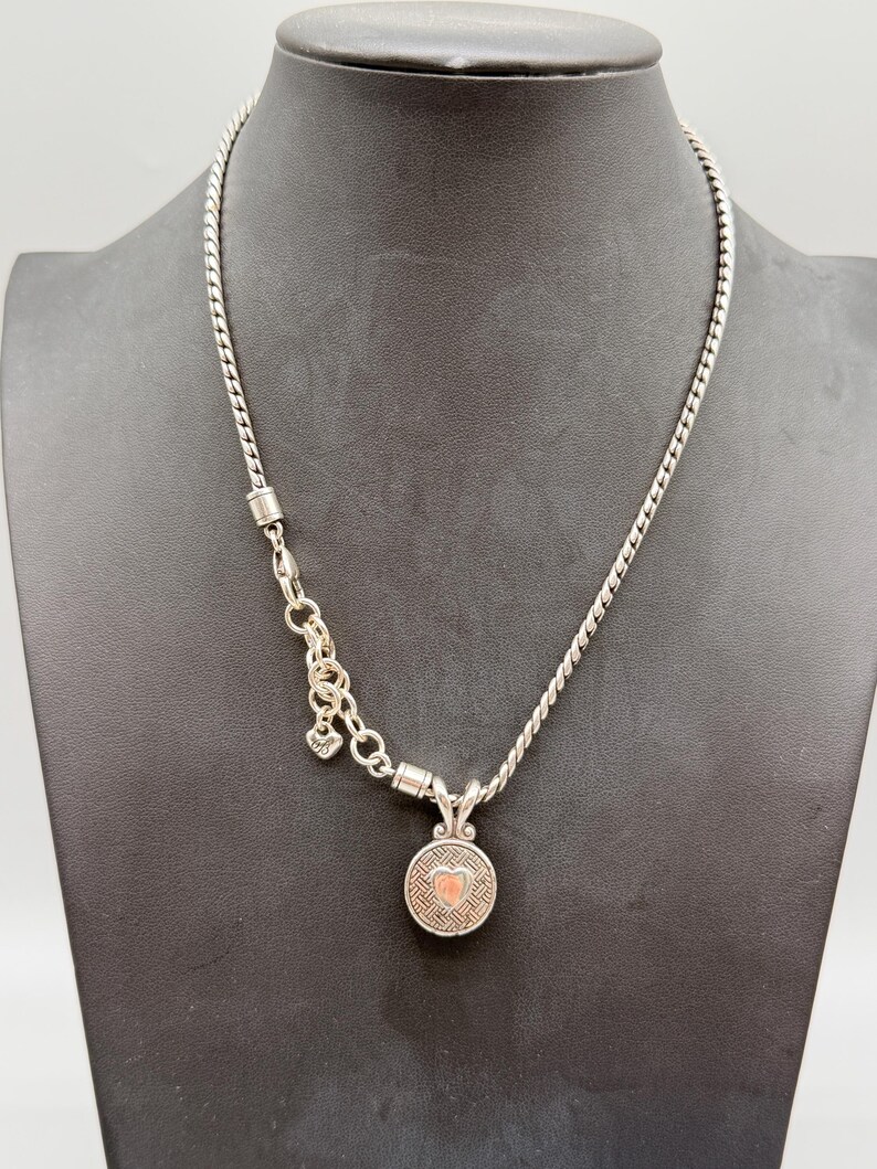 May include: A silver-toned necklace featuring a twisted chain and a circular pendant with a textured geometric pattern and a small heart-shaped center. The necklace includes a short section of larger link chain with a small dangling charm.