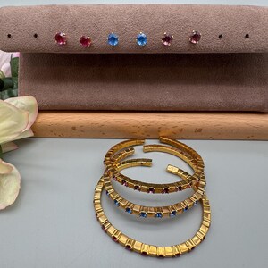 May include: Gold-toned bracelets and stud earrings. The bracelets are adorned with rows of red and blue gemstones. The earrings are displayed on a brown velvet surface and feature red, blue, and purple gemstones.