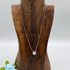 May include: A silver necklace with a small, square pendant featuring a cluster of sparkling stones. The necklace is displayed on a wooden jewelry bust against a white background. A small bouquet of blue flowers is in the lower left corner.