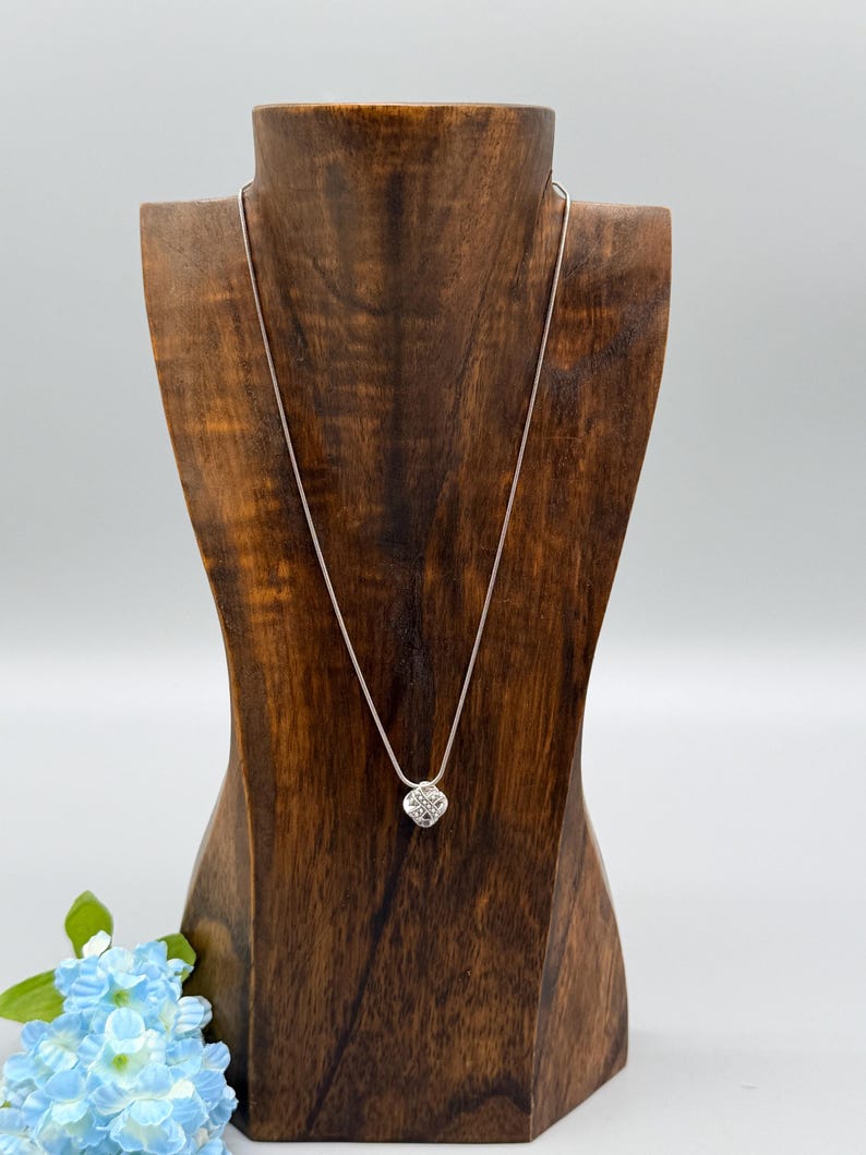 May include: A silver necklace with a small, square pendant featuring a cluster of sparkling stones. The necklace is displayed on a wooden jewelry bust against a white background. A small bouquet of blue flowers is in the lower left corner.