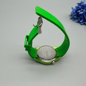 May include: A bright lime green wristwatch with a flexible silicone strap and a silver-toned metal buckle. The circular watch case back is made of stainless steel and features the engraved text IZOD ESSENTIAL in the center of the casing.