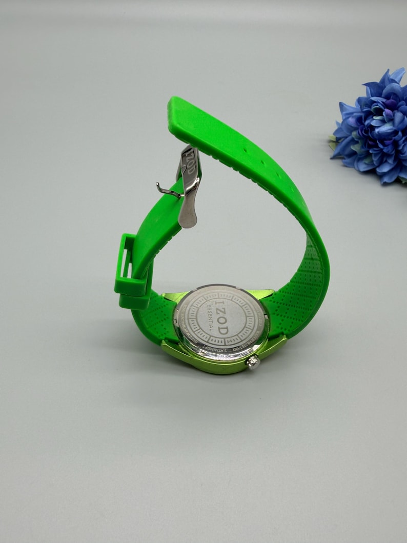 May include: A bright lime green wristwatch with a flexible silicone strap and a silver-toned metal buckle. The circular watch case back is made of stainless steel and features the engraved text IZOD ESSENTIAL in the center of the casing.
