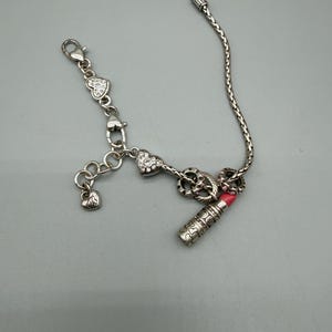 May include: A silver-toned charm bracelet featuring a braided chain, a lobster clasp, and several dangling charms. The charms include heart shapes with embedded clear crystals, a small plain heart, and a detailed lipstick tube with a pink tip.