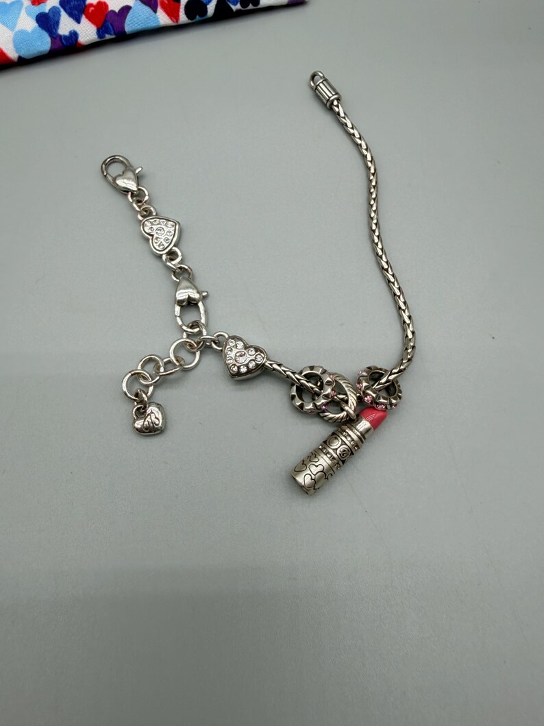 May include: A silver-toned charm bracelet featuring a braided chain, a lobster clasp, and several dangling charms. The charms include heart shapes with embedded clear crystals, a small plain heart, and a detailed lipstick tube with a pink tip.