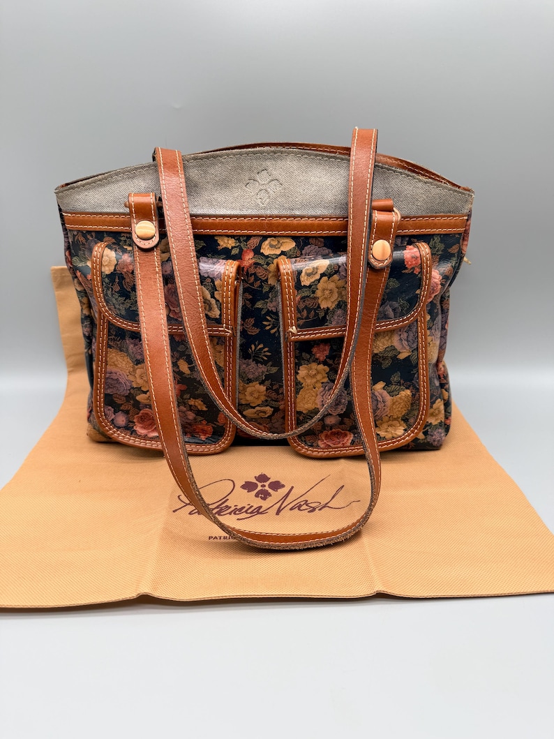 May include: A floral print tote bag with brown leather handles and accents. The bag features a dark background with colorful flowers and multiple exterior pockets. The brand name "Patricia Nash" is visible on the bag's dust cover.