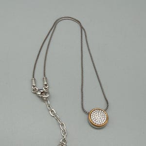 Puede incluir: Collar plateado con un colgante redondo. El colgante tiene un borde dorado y est&aacute; lleno de peque&ntilde;os cristales transparentes. El collar tiene una cadena ajustable con un dije en forma de coraz&oacute;n.