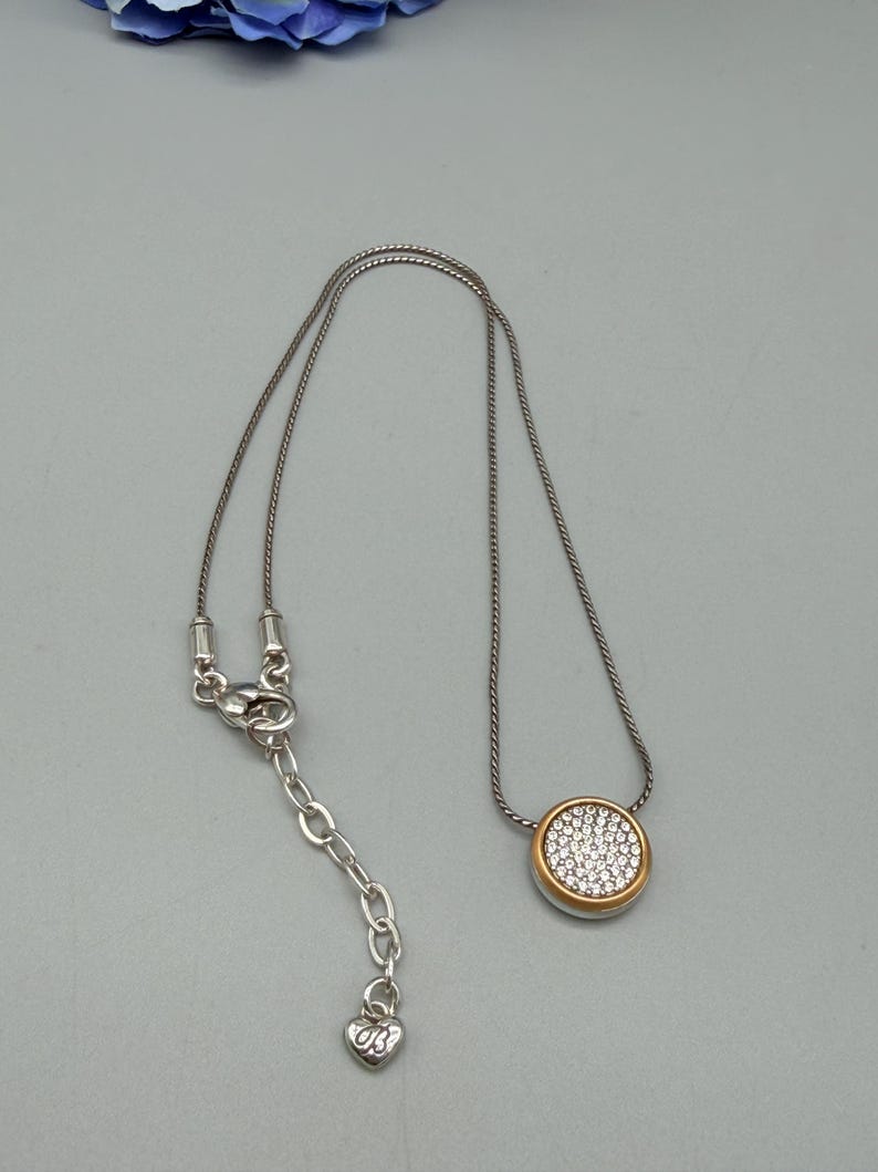 Puede incluir: Collar plateado con un colgante redondo. El colgante tiene un borde dorado y est&aacute; lleno de peque&ntilde;os cristales transparentes. El collar tiene una cadena ajustable con un dije en forma de coraz&oacute;n.