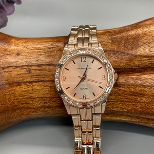 May include: A rose gold wristwatch with a round face and a metal band. The watch face has a white background with black Roman numeral hour markers and a quartz movement. The watch is surrounded by a bezel with clear rhinestones.