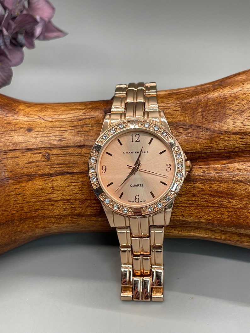 May include: A rose gold wristwatch with a round face and a metal band. The watch face has a white background with black Roman numeral hour markers and a quartz movement. The watch is surrounded by a bezel with clear rhinestones.