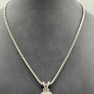 May include: A silver-toned necklace featuring a twisted rope chain and a circular pendant. The pendant has a central clear stone surrounded by a gold-toned inner ring and a silver-toned twisted rope border, suspended from a decorative bail.
