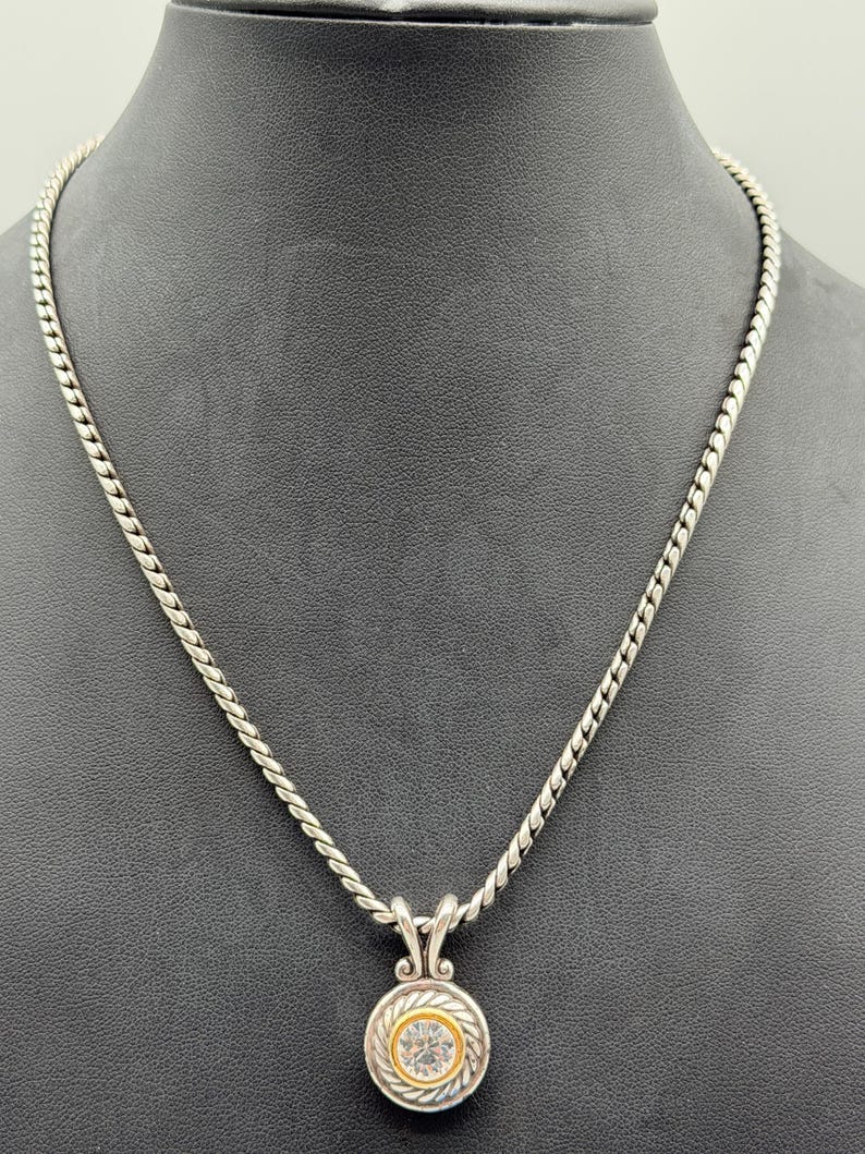 May include: A silver-toned necklace featuring a twisted rope chain and a circular pendant. The pendant has a central clear stone surrounded by a gold-toned inner ring and a silver-toned twisted rope border, suspended from a decorative bail.