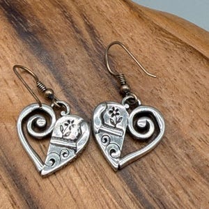 May include: A pair of silver heart-shaped earrings. Each earring has a split heart design with a floral accent and scrollwork. The earrings have a hook closure and are displayed on a wooden surface.