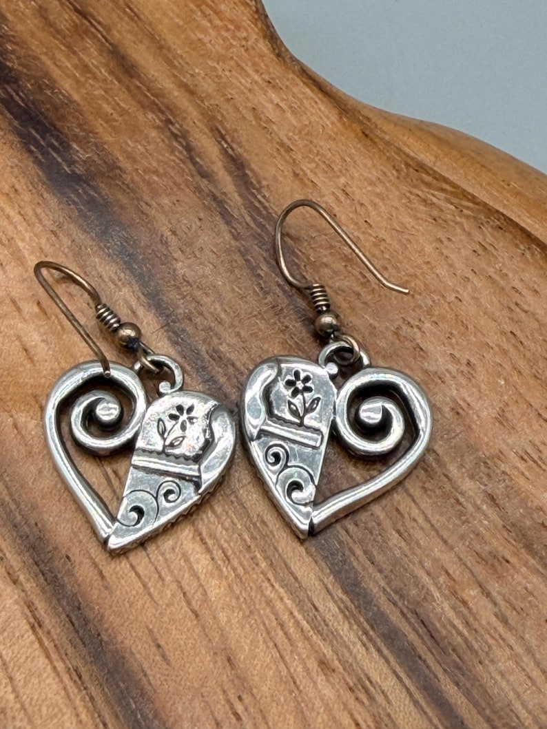 May include: A pair of silver heart-shaped earrings. Each earring has a split heart design with a floral accent and scrollwork. The earrings have a hook closure and are displayed on a wooden surface.