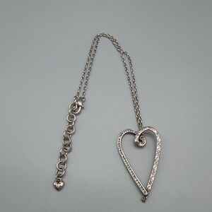 Pu&ograve; includere: Collana con pendente a cuore argentato. Il design a cuore aperto &egrave; impreziosito da piccole pietre trasparenti e volute decorative. La collana ha una catena regolabile con un ciondolo a forma di cuore all'estremit&agrave;.