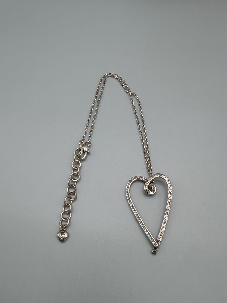 Pu&ograve; includere: Collana con pendente a cuore argentato. Il design a cuore aperto &egrave; impreziosito da piccole pietre trasparenti e volute decorative. La collana ha una catena regolabile con un ciondolo a forma di cuore all'estremit&agrave;.