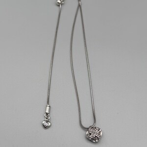 May include: A silver-toned necklace with a small, round pendant featuring a woven design. The necklace has a delicate chain and an adjustable clasp. A heart-shaped charm with the initials "DB" hangs near the clasp.