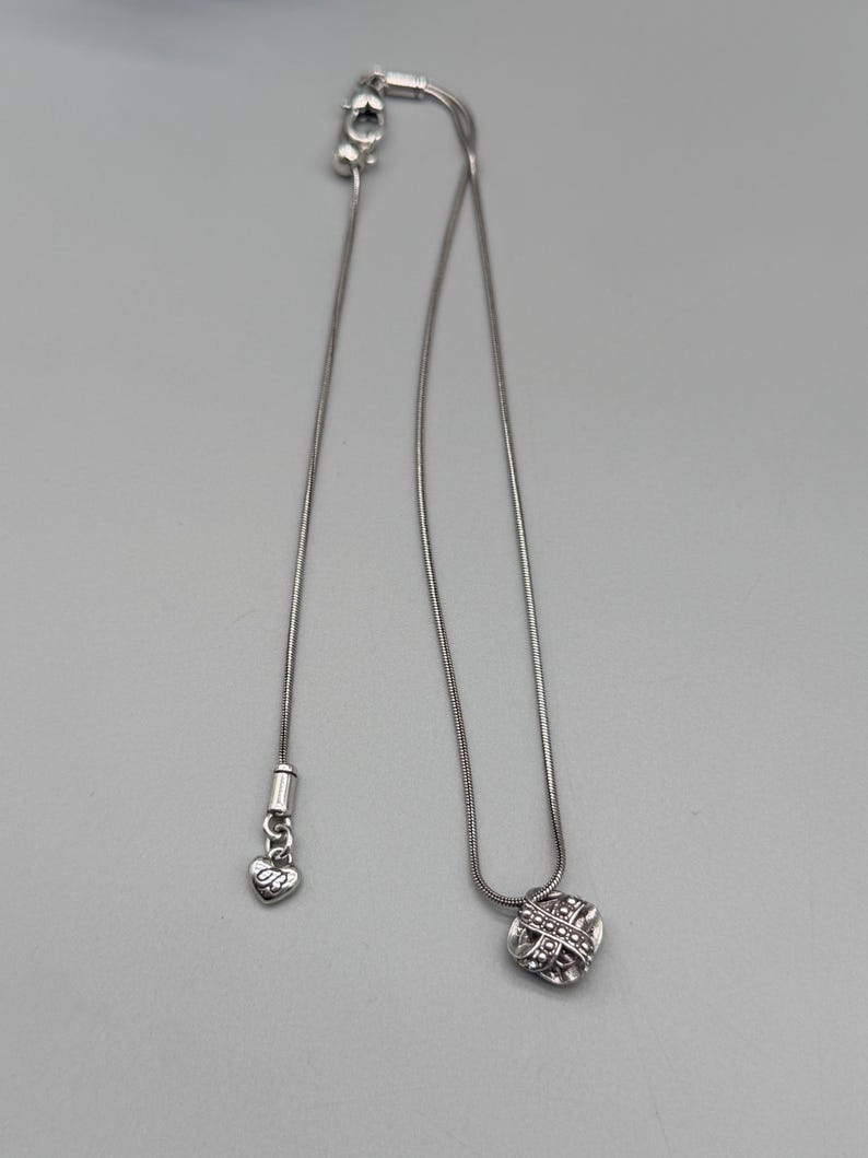 May include: A silver-toned necklace with a small, round pendant featuring a woven design. The necklace has a delicate chain and an adjustable clasp. A heart-shaped charm with the initials "DB" hangs near the clasp.