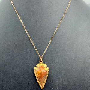 May include: A gold-toned chain necklace featuring an arrowhead-shaped pendant. The pendant is crafted from a translucent, multi-colored stone with streaks of orange, brown, and red, encased in a textured, metallic gold-colored frame.