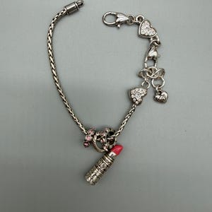 May include: A silver-toned charm bracelet featuring a braided chain, a lipstick charm with a pink tip, a ring charm with pink stones, and multiple heart-shaped charms adorned with clear crystals, secured with a lobster claw clasp closure.
