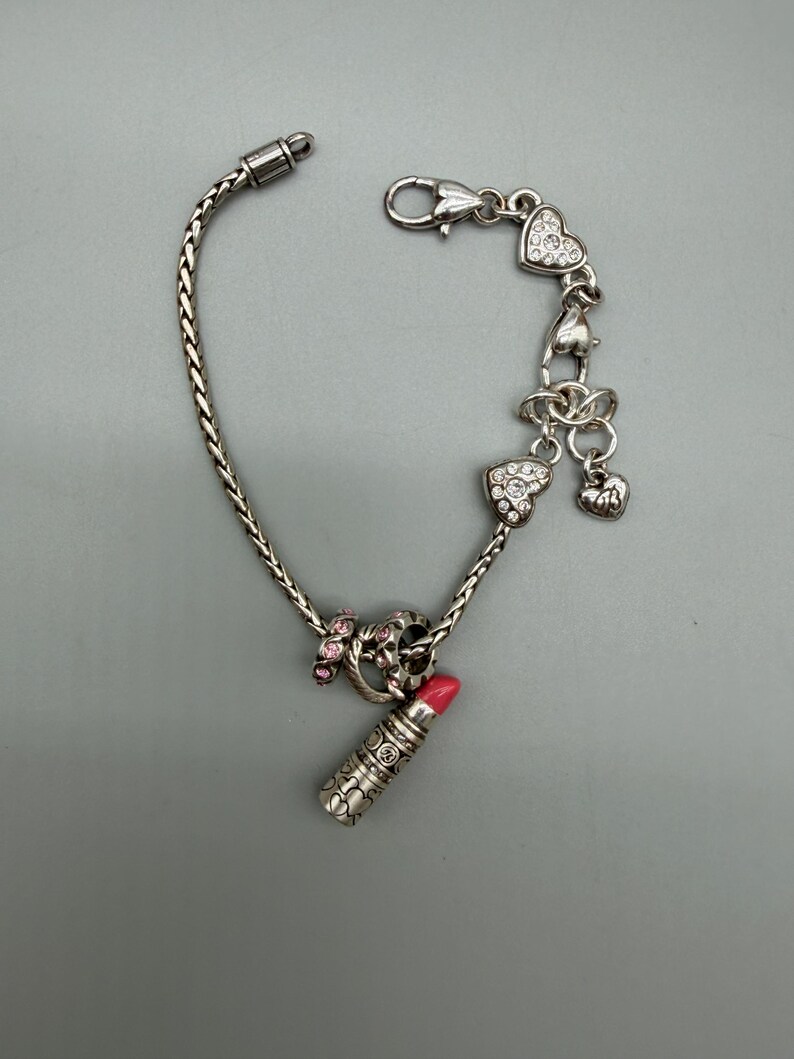 May include: A silver-toned charm bracelet featuring a braided chain, a lipstick charm with a pink tip, a ring charm with pink stones, and multiple heart-shaped charms adorned with clear crystals, secured with a lobster claw clasp closure.