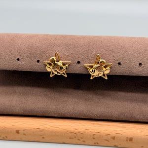 May include: A pair of gold-colored star-shaped stud earrings. Each earring features a Mickey Mouse design in the center of the star. The earrings are displayed on a brown surface with a wooden base.