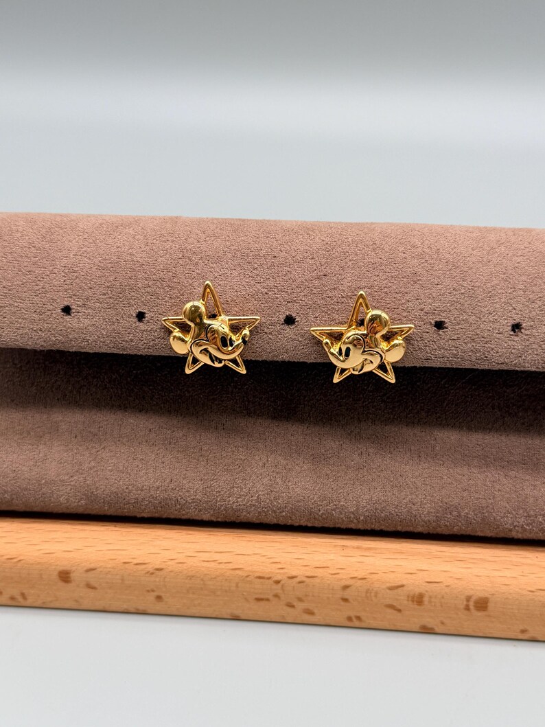 May include: A pair of gold-colored star-shaped stud earrings. Each earring features a Mickey Mouse design in the center of the star. The earrings are displayed on a brown surface with a wooden base.