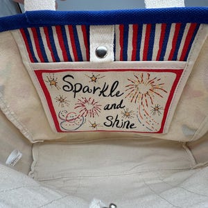 Puede incluir: Un bolso de mano de lona con un borde superior azul y asas blancas. El interior presenta un bolsillo con rayas rojas, blancas y azules y las palabras "Sparkle and Shine" con ilustraciones de fuegos artificiales.