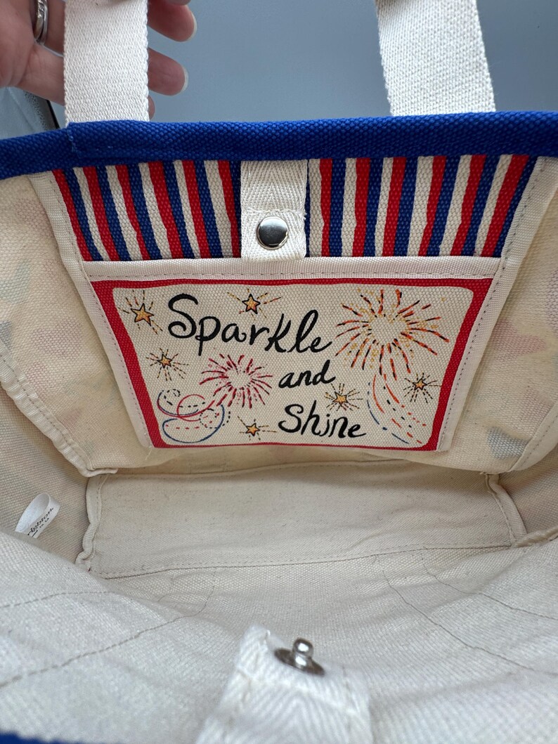 Puede incluir: Un bolso de mano de lona con un borde superior azul y asas blancas. El interior presenta un bolsillo con rayas rojas, blancas y azules y las palabras "Sparkle and Shine" con ilustraciones de fuegos artificiales.