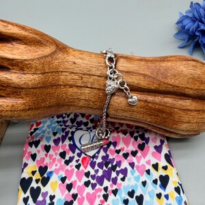 May include: A silver-toned charm bracelet featuring a heart-shaped charm with embedded clear stones, a small plain heart charm, and a lipstick-shaped charm. The bracelet has a woven chain design and is displayed on a wooden stand.