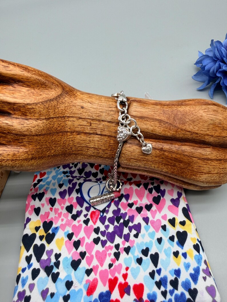 May include: A silver-toned charm bracelet featuring a heart-shaped charm with embedded clear stones, a small plain heart charm, and a lipstick-shaped charm. The bracelet has a woven chain design and is displayed on a wooden stand.