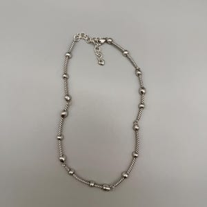 May include: A silver-toned necklace featuring a twisted rope chain design interspersed with smooth, round metallic beads. The necklace includes an adjustable chain section at the clasp, which is finished with a small, heart-shaped charm.