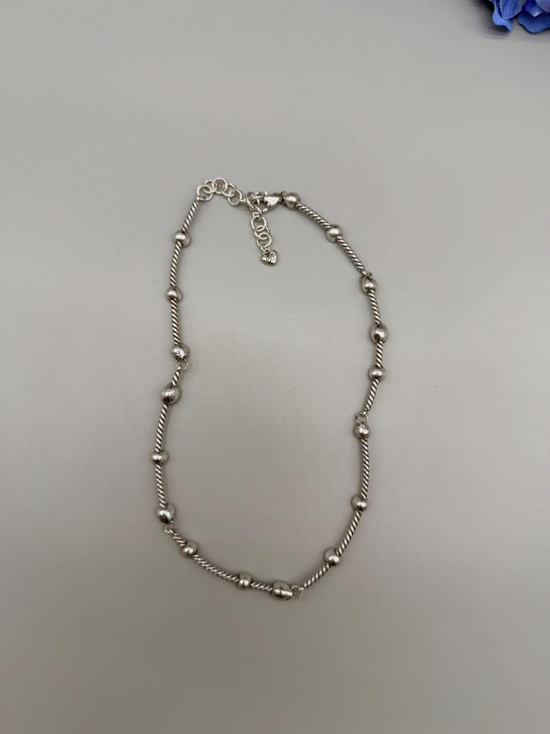 May include: A silver-toned necklace featuring a twisted rope chain design interspersed with smooth, round metallic beads. The necklace includes an adjustable chain section at the clasp, which is finished with a small, heart-shaped charm.
