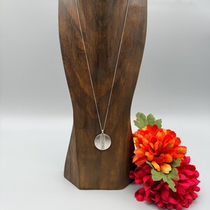 Puede incluir: Un collar con colgante plateado con un colgante redondo y texturizado se exhibe en un expositor de joyas de madera. El collar cuelga de una delicada cadena. Un ramo de flores rojas y naranjas se coloca a la derecha del expositor.