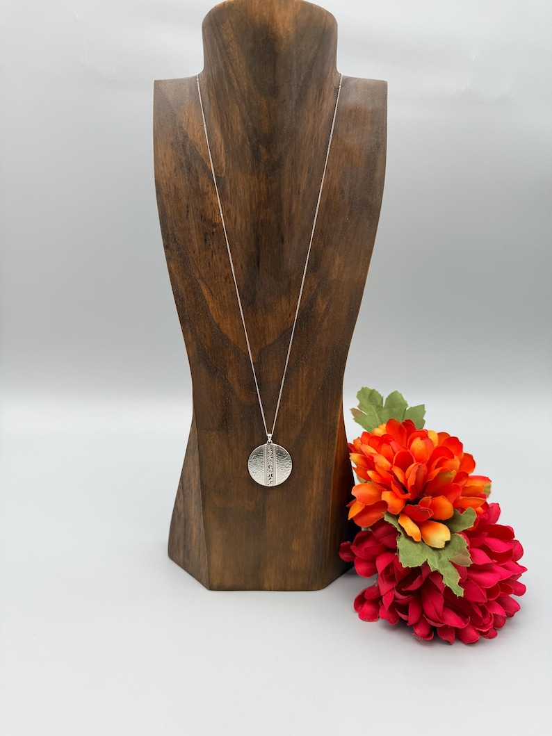 Puede incluir: Un collar con colgante plateado con un colgante redondo y texturizado se exhibe en un expositor de joyas de madera. El collar cuelga de una delicada cadena. Un ramo de flores rojas y naranjas se coloca a la derecha del expositor.