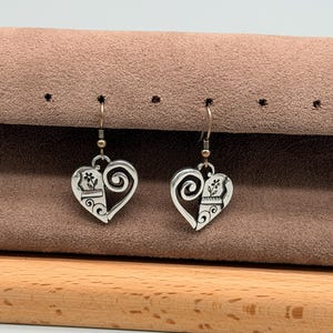 May include: A pair of silver-toned heart-shaped earrings. Each earring features a floral design and a swirling accent. The earrings hang from a simple hook. The earrings are displayed on a brown surface.