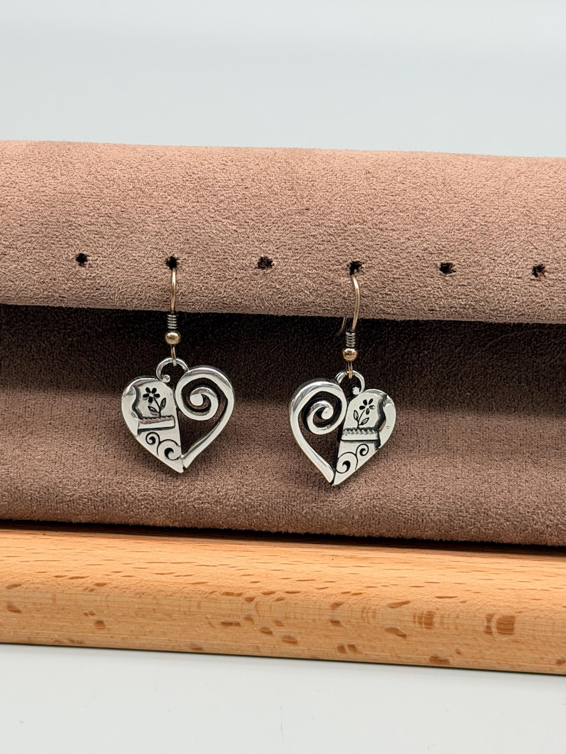 May include: A pair of silver-toned heart-shaped earrings. Each earring features a floral design and a swirling accent. The earrings hang from a simple hook. The earrings are displayed on a brown surface.