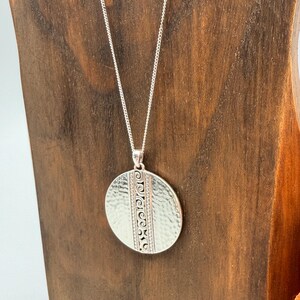 Puede incluir: Un collar con colgante de plata con textura martillada y detalles decorativos grabados. El colgante ovalado cuelga de una delicada cadena de plata. El collar se exhibe sobre un soporte de madera.