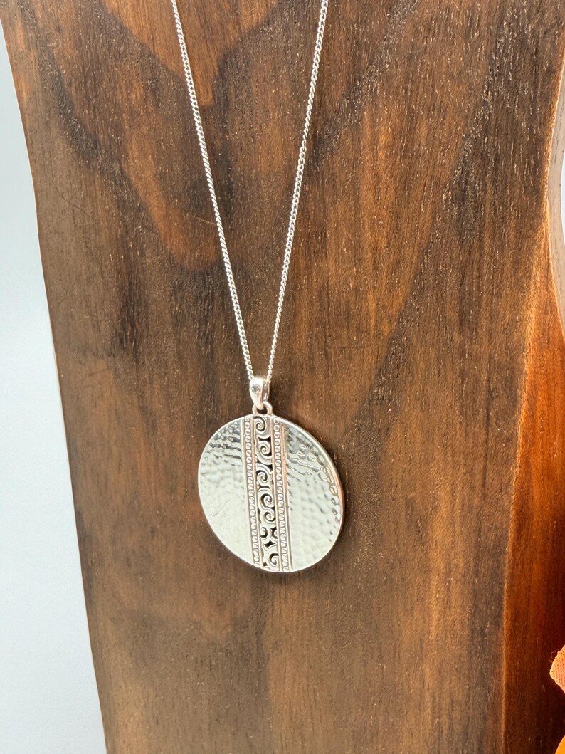 Puede incluir: Un collar con colgante de plata con textura martillada y detalles decorativos grabados. El colgante ovalado cuelga de una delicada cadena de plata. El collar se exhibe sobre un soporte de madera.