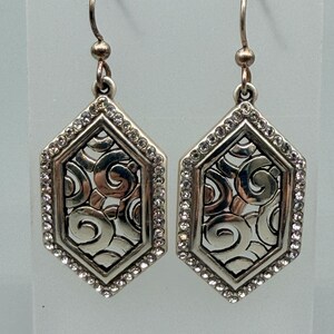 Puede incluir: Un par de pendientes plateados. Cada pendiente presenta una forma geom&eacute;trica hexagonal con un dise&ntilde;o decorativo de espirales en el centro. El borde exterior est&aacute; bordeado con peque&ntilde;os cristales transparentes. Los pendientes tienen un cierre de gancho.