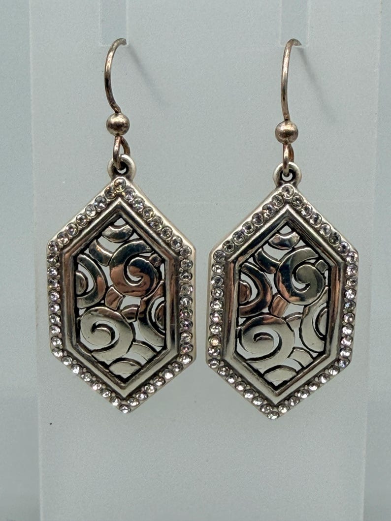 Puede incluir: Un par de pendientes plateados. Cada pendiente presenta una forma geom&eacute;trica hexagonal con un dise&ntilde;o decorativo de espirales en el centro. El borde exterior est&aacute; bordeado con peque&ntilde;os cristales transparentes. Los pendientes tienen un cierre de gancho.