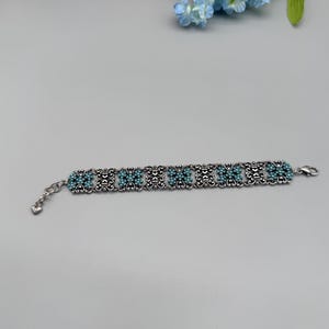 以下が含まれることがあります： ターコイズビーズと繊細な透かし模様が施されたシルバーカラーのブレスレット。ブレスレットは、装飾的な金属細工の中にセットされたターコイズビーズのクラスターが特徴です。留め金はロブスタークラスプスタイルです。ブレスレットは、明るい灰色の表面に表示されています。