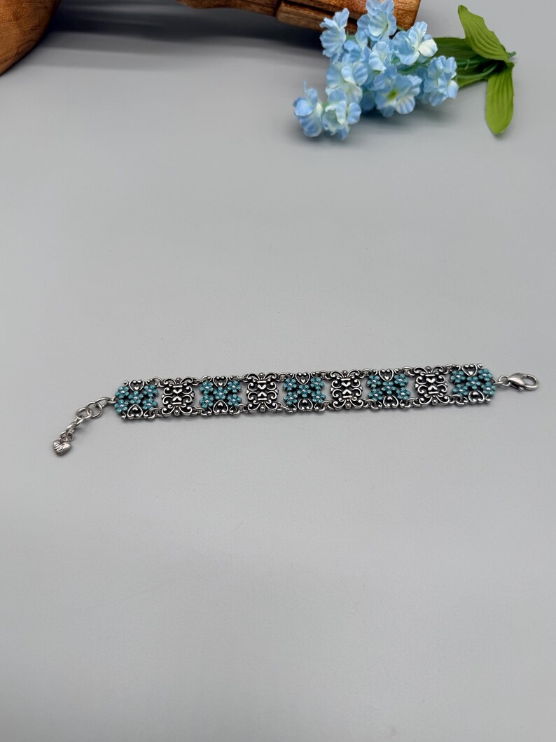 以下が含まれることがあります： ターコイズビーズと繊細な透かし模様が施されたシルバーカラーのブレスレット。ブレスレットは、装飾的な金属細工の中にセットされたターコイズビーズのクラスターが特徴です。留め金はロブスタークラスプスタイルです。ブレスレットは、明るい灰色の表面に表示されています。