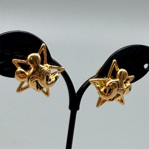 May include: A pair of gold-colored Mickey Mouse earrings. Each earring features a Mickey Mouse figure within a star-shaped frame. The earrings are displayed on a black earring stand against a light gray background. The earrings are likely made of metal.