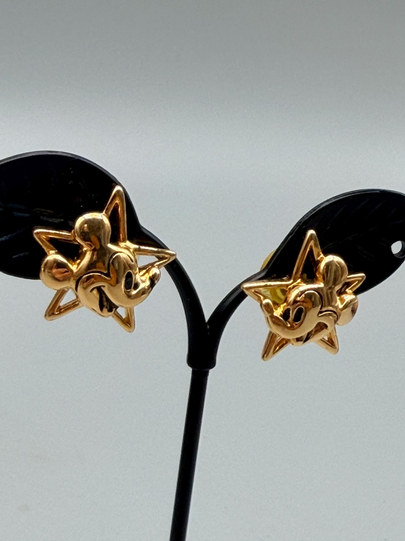 May include: A pair of gold-colored Mickey Mouse earrings. Each earring features a Mickey Mouse figure within a star-shaped frame. The earrings are displayed on a black earring stand against a light gray background. The earrings are likely made of metal.
