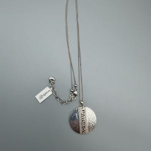 Puede incluir: Collar Brighton plateado con un colgante redondo. El colgante tiene una textura martillada en un lado y un patr&oacute;n decorativo en el otro. El collar incluye una cadena y una etiqueta que dice "Brighton."