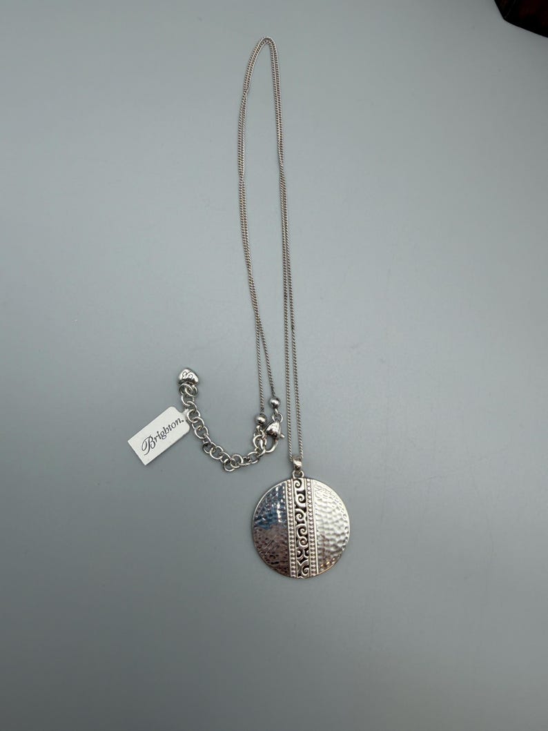Puede incluir: Collar Brighton plateado con un colgante redondo. El colgante tiene una textura martillada en un lado y un patr&oacute;n decorativo en el otro. El collar incluye una cadena y una etiqueta que dice "Brighton."