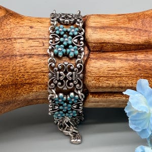 以下が含まれることがあります： ターコイズブルーの花のアクセントが付いた、装飾的なデザインのシルバーカラーのブレスレット。 ブレスレットは、透かし模様と花のディテールが施された複数の長方形のリンクで構成されています。 クラスプは装飾的なフックとアイクロージャーです。