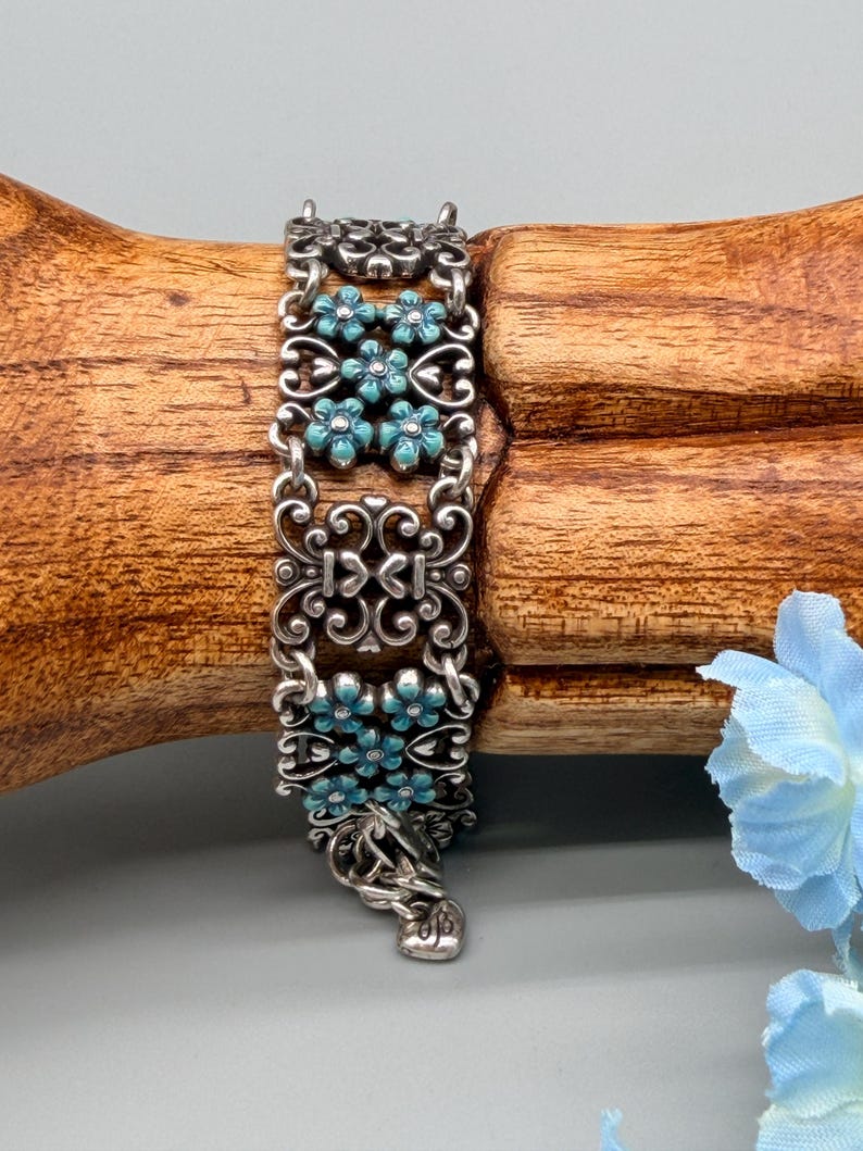 以下が含まれることがあります： ターコイズブルーの花のアクセントが付いた、装飾的なデザインのシルバーカラーのブレスレット。 ブレスレットは、透かし模様と花のディテールが施された複数の長方形のリンクで構成されています。 クラスプは装飾的なフックとアイクロージャーです。