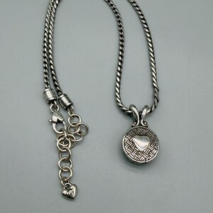 May include: A silver-toned necklace featuring a round pendant with a textured, woven-style pattern and a smooth, raised heart in the center. The chain is a thick, twisted rope style with a lobster claw clasp and an adjustable link extender.